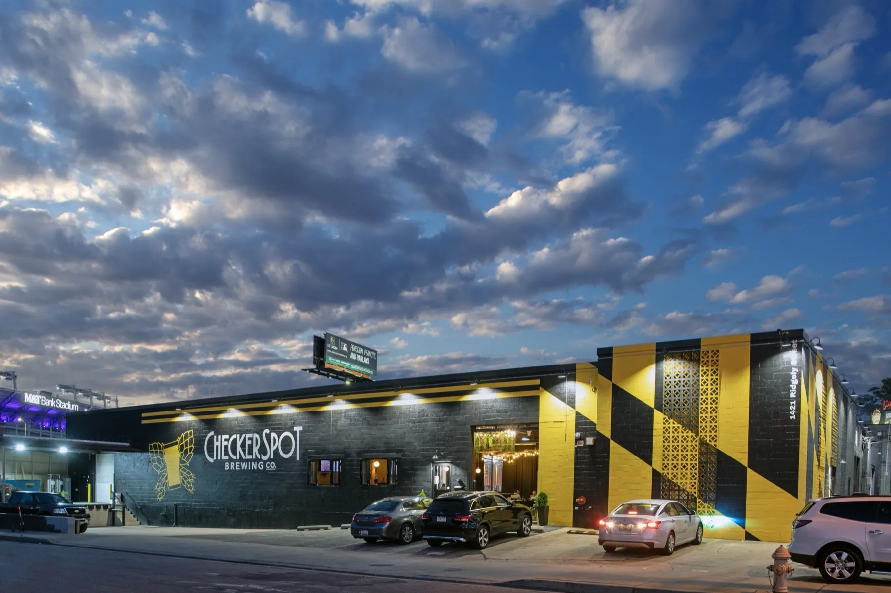 The Checkerspot Brewing building at dusk on Ridgely Street, its Maryland-flag corner lit, with M&T Bank Stadium behind it