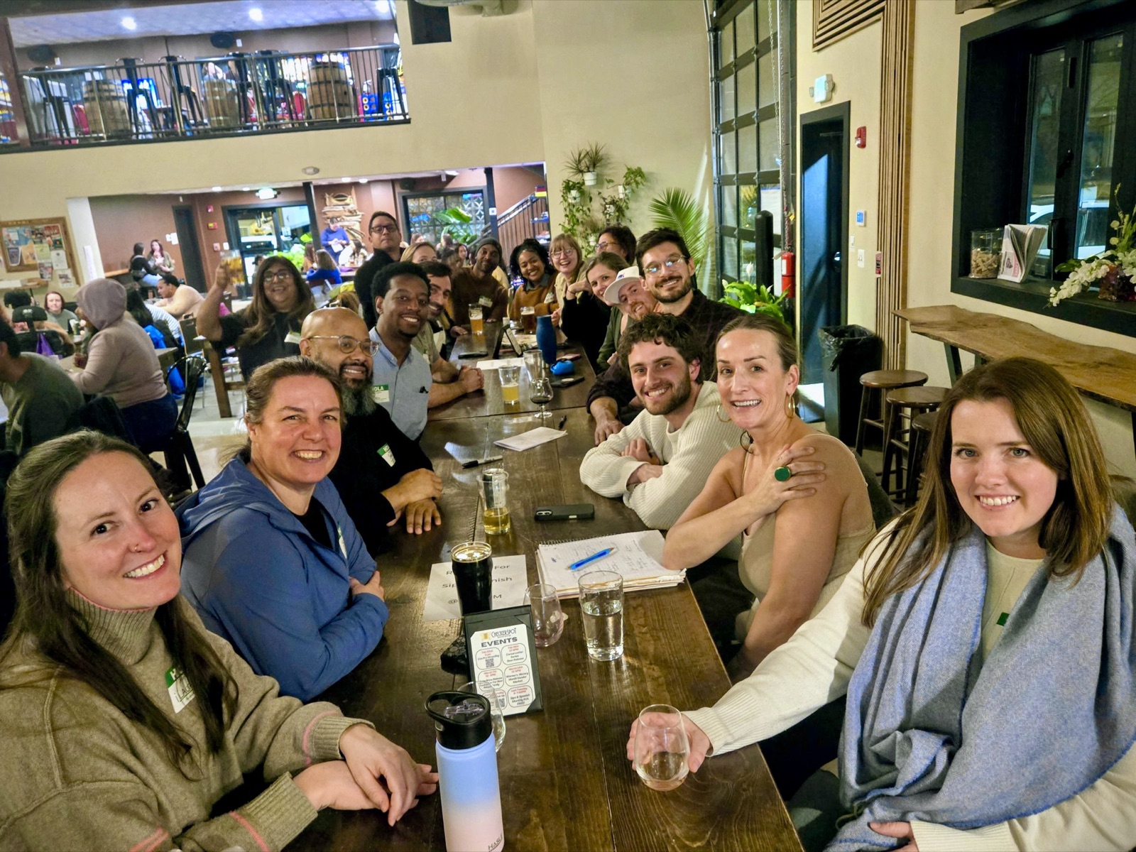 A long table of smiling friends and regulars sharing beers together in the Checkerspot Brewing taproom in Baltimore